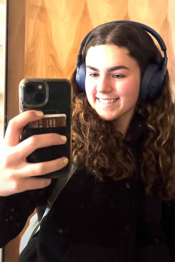 Anna Godlewski, a female student smiles into her mirror while holding up her cell phone to record a video. She has long brown hair and is wearing a pair of headphones.